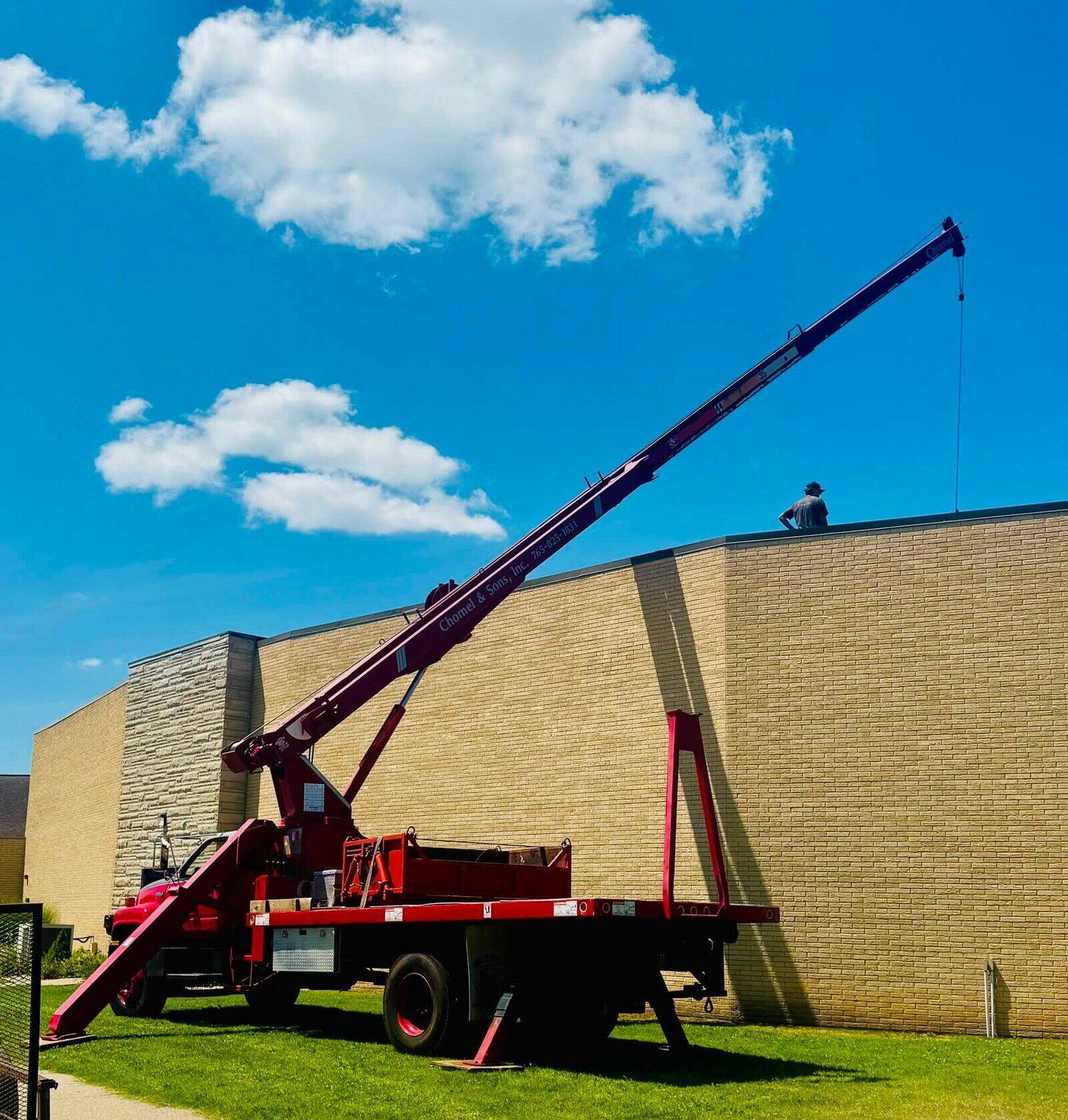 A Chomel and Sons red boom truck lifting roofing material to a roof.